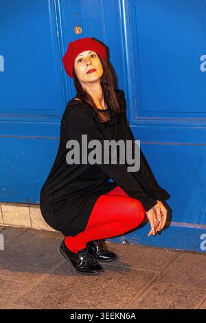 Frau mit roter Baskenmütze und Strumpfhosen, die sich an der blauen Tür hockt Stockfoto