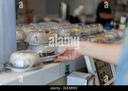 Nahaufnahme einer Hand eines Kunden, die eine frische Sushi-Platte aus einem beweglichen Förderband in einem Restaurant in Brisbane auswählt. (2025) Stockfoto