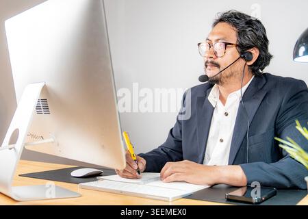 Ein junger männlicher Mitarbeiter kommuniziert mit Kollegen über ein virtuelles Meeting auf seinem Laptop in einer modernen Büroumgebung. Stockfoto