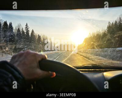 Ein Fahrer blickt von der Windschutzscheibe auf die Straße vor sich, während die Sonne während eines Reiseabenteuers im Hintergrund untergeht. Verschwommenes Bild Stockfoto