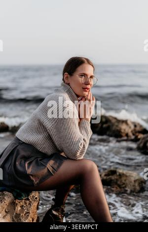 Eine Frau mit Ozeanpullover, die an der felsigen Küste sitzt, Brille und stilvolle Strickwaren trägt und bei Sonnenuntergang nachdenklich an den Wellen aussieht Stockfoto