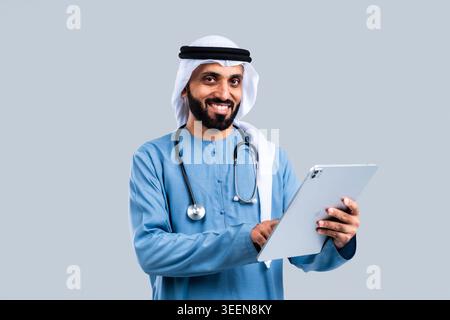 Emirati männlicher Arzt trägt traditionelle Kleidung und ein Stethoskop, lächelt, während er ein digitales Tablet hält, repräsentiert Telehealth, moderne Gesundheitsversorgung Stockfoto