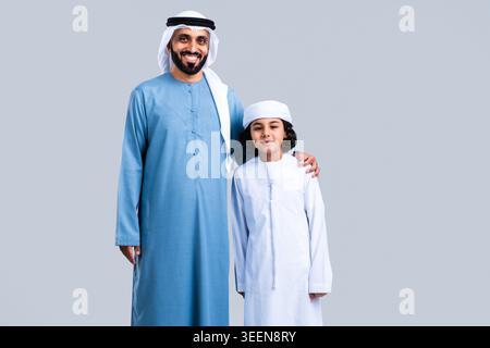 Der arabische Vater lächelt, trägt traditionelle Kandora und Gghutra, Arm um seinen Sohn, der auch traditionelle Kleidung trägt, und zeigt Familienbande und stolzen Vater Stockfoto