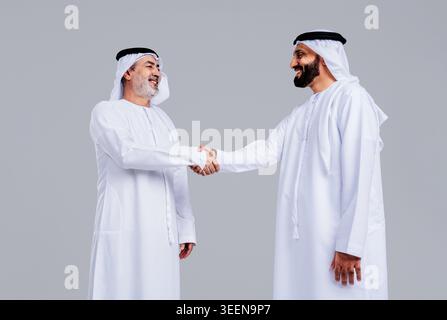 Zwei lächelnde arabische Geschäftsmänner, in traditioneller Kleidung gekleidet, Hände schütteln, was eine erfolgreiche Partnerschaft, Vereinbarung, Vertrauen, Zusammenarbeit bedeutet Stockfoto
