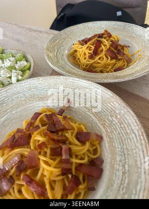 Hausgemachte Spaghetti Carbonara mit knusprigem Speck, serviert neben frischem Gurken- und Fetasalat in Keramikschüsseln. Stockfoto