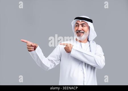 Älterer emirati in traditioneller Kandora und Gghutra lächelnd und mit Händen und Fingern zur Seite zeigend, selbstbewusst und einladend auf grauem Studio b Stockfoto