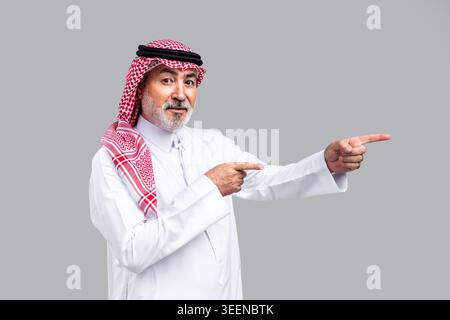 Saudischer Seniorenmann, der traditionelle Gghutra und Thobe trägt, selbstbewusst lächelt und nach rechts zeigt, die Richtung zeigt oder wichtige Informationen hervorhebt Stockfoto