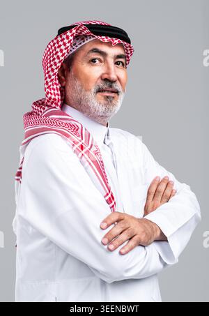 Saudischer Seniorenmann mit traditionellem Thobe und Gghutra mit Agar, sich kreuzenden Armen, während er den Betrachter auf einem neutralen grauen Hintergrund selbstbewusst anlächelt Stockfoto