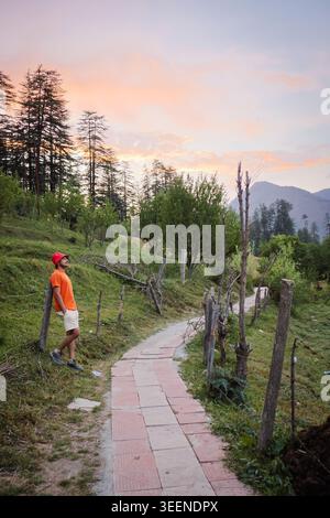 Ein Mann in einem orangefarbenen Hemd und einer roten Mütze beugt sich an einen Holzzaun neben einem gepflasterten Weg. Die Szene spielt in einer hügeligen Landschaft mit Bäumen und Bergen Stockfoto