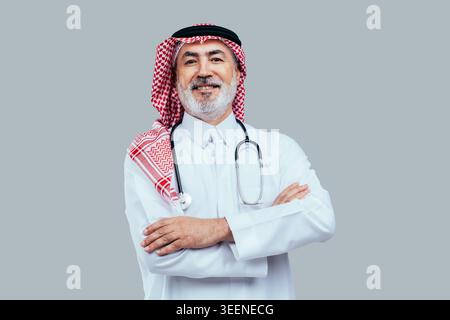 Senior saudi-arabischer Arzt in traditionellem Dschungel und Gghutra mit Stethoskop, Arme überkreuzt und lächelnd vor grauem Hintergrund, selbstbewusst erfahren Stockfoto