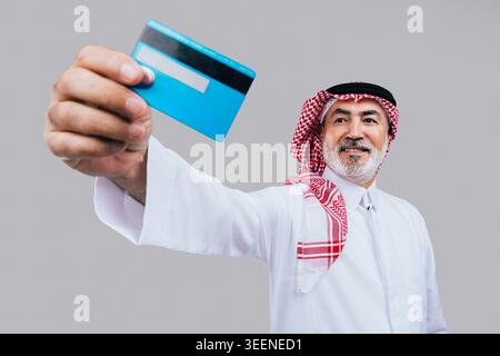 Saudischer Seniorenmann mit traditionellem Gghutra und Thobe, lächelnd, während er eine Kreditkarte hält, symbolisiert modernes Bankwesen, E-Commerce und finanztransaktionen Stockfoto