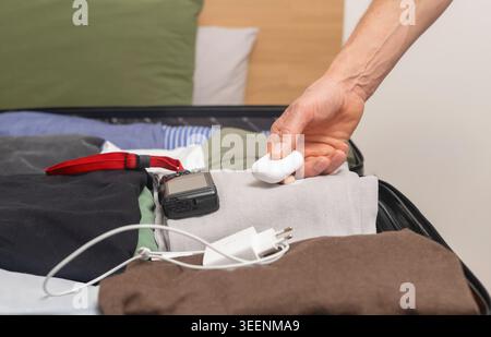 Mann, der das Ladegerät und das Kabel in einen offenen Koffer mit gefalteter Kleidung legt. Erwachsene männliche Hände, die elektronisches Zubehör im Gepäck arrangieren. Reisevorbereitung Stockfoto