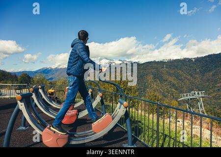 Ein Junge steht auf Fitnessgeräten in einem Fitnessbereich im Freien. Er sieht schneebedeckte Berge unter blauem Himmel. Im Fitnessbereich sind Bäume zu sehen. Stockfoto