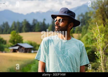 Ein bärtiger Mann mit gemustertem T-Shirt, Hut und Sonnenbrille schaut auf die Berglandschaft in Shangarh, Himachal Pradesh, Indien. Stockfoto