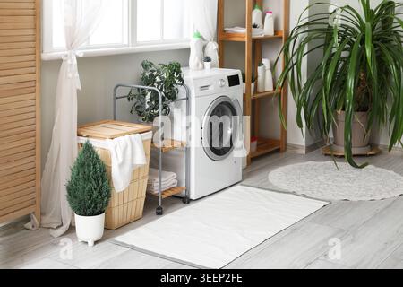 Waschmaschine, Wäschekorb und Klappschirm neben dem Fenster im Zimmer Stockfoto