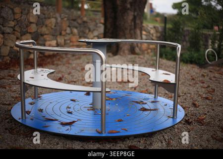 Leeres blaues Metall-Karussell auf einem Spielplatz, der mit Herbstlaub bedeckt ist. Aufgelassenes Kinderkonzept in saisonaler Umgebung. Stockfoto
