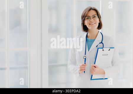 Schöne lächelnde Praktikant hält Zwischenablage mit schriftlichen Papieren und Stift, trägt weiße Uniform, bereit, Patienten in eigener Privatklinik zu sehen, steht innen vor weißem Hintergrund. Bereich für Kopierfläche Stockfoto