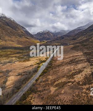 Aus der Vogelperspektive auf die A82, die sich durch das dramatische Glen Coe Tal schlängelt, umgeben von schneebedeckten Bergen und braunem Moor unter einem bewölkten Himmel Stockfoto