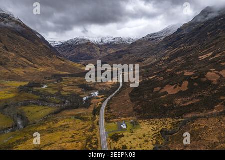 Aus der Vogelperspektive auf die gewundene A82-Straße durch das dramatische Glencoe-Tal mit schneebedeckten Bergen und einem kleinen weißen Häuschen unter einem stimmungsvollen Himmel Scotlan Stockfoto