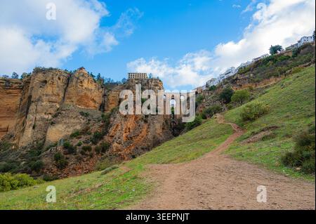Die Steinbrücke Puente Nuevo überspannt eine tiefe Schlucht zwischen felsigen Klippen in Ronda, Andalusien, Spanien. Ein Schotterweg führt einen grasbewachsenen Hügel hinauf in Richtung weißes Buil Stockfoto