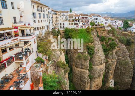 Weiße Wohnhäuser und Restaurantterrassen stehen am Rande einer steilen, felsigen Schlucht, die mit grüner Vegetation bedeckt ist, in Ronda, Andalusien, Spanien. Stockfoto