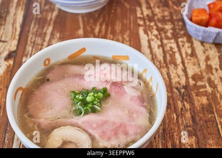 Eine warme Schüssel mit Hühnerbrühe, garniert mit zartem Chashu und grünen Zwiebeln, mit einer Nahaufnahme welliger Nudeln auf Essstäbchen auf einem Holztisch. Stockfoto