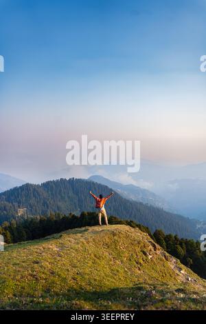 Ein Wanderer mit Rucksack steht auf einem grasbewachsenen Bergrücken mit Blick auf ein Tal im Raghupur Fort, Sojha, Manali, Himachal Pradesh, Indien. Stockfoto