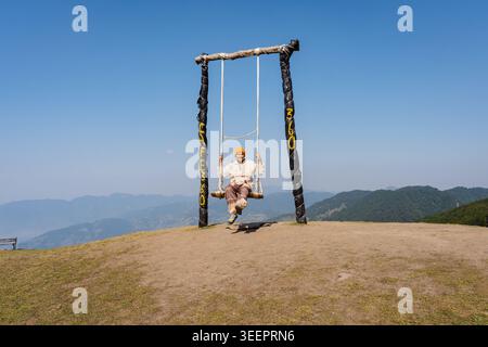 Ein Mann sitzt auf einer rustikalen Holzschaukel auf einem Hügel mit Blick auf die Berge im Raghupur Fort, Sojha, Manali, Himachal Pradesh, Indien. Stockfoto
