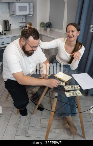 Junge glückliche Paare, die monatliche Ausgaben und Einkommen berechnen, Dollar-Banknoten zählen und Zahlen an einem Glastisch in ihrer Küche aufschreiben, planen Stockfoto