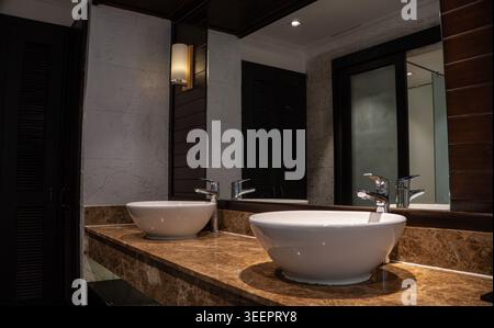 Elegantes Doppelgefäß mit Waschbecken auf einer braunen Marmorarbeitsfläche in einem luxuriösen Hotelbad in Vietnam. Chrom-Armaturen, Wandleuchte, großer Spiegel und dunkler wo Stockfoto