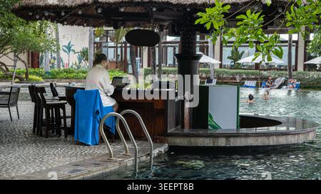 Frau in einem weißen Bademantel, die an einem Laptop an einer Poolbar in einem tropischen Resort in Vietnam arbeitet. Poolbar, Strohdach, Schwimmer im Hintergrund, blauer Schlepper Stockfoto