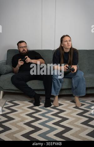 Junge Paare, die auf einer Couch sitzen, Gamecontroller halten und sich intensiv konzentrieren, während sie gemeinsam im Wohnzimmer Videospiele spielen und dabei LE genießen Stockfoto