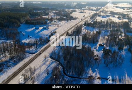 Aus der Vogelperspektive einer geraden Autobahn, die durch schneebedeckte Felder und dichte Wälder unter hellem Wintersonnenlicht in Litauen führt. Stockfoto