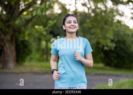 Junge indianerin fühlt sich beim Laufen im Freien positiv und gesund, trägt Sportbekleidung und eine Smartwatch und konzentriert sich auf ihre Fitnessziele in einer grünen Parkumgebung Stockfoto