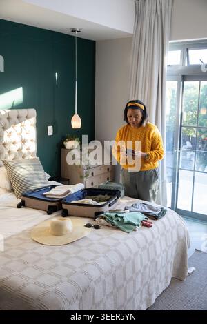 Reife Frau mit gelbem Pullover, die offenen Koffer mit Telefon im Schlafzimmer, Kopierraum packt Stockfoto