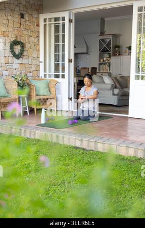Ältere asiatische Frau kniend auf grüner Terrassenmatte, um Uhr in Top dunklen Leggings in der Nähe von Kurzhanteln zu sehen Stockfoto