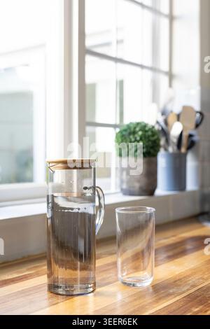 Pitcher und Glas befinden sich auf einer sonnenbeleuchteten Arbeitsplatte durch ein Mehrscheibenfenster mit Pflanzen- und Löffelhalter Stockfoto