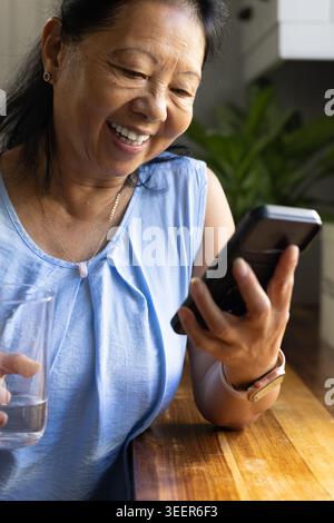 Ältere asiatische Frau lächelt, sieht auf das Telefon, hält Glas in blauer Bluse am Tisch in der Küche Stockfoto