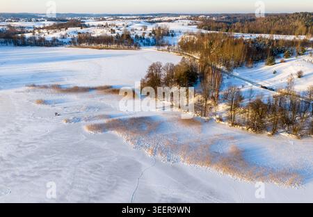 Aus der Vogelperspektive auf einen gefrorenen See mit Schilf und ein von Bäumen gesäumtes Ufer unter goldenem Sonnenlicht in Litauen. Stockfoto