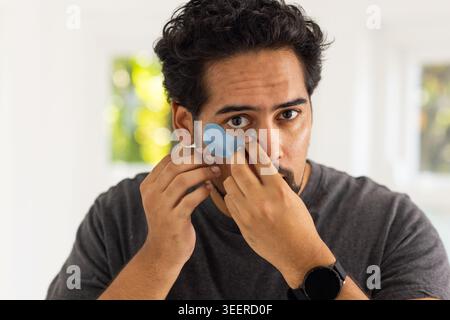 Mittelerwachsener Mann, der blaue sichelförmige Hydrogel unter den Augen aufträgt, zu Hause, mit grauem T-Shirt Stockfoto