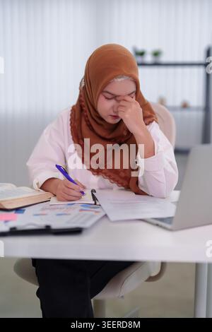 Gestresste junge asiatische Muslimfrau mit Hijab, die Kopfschmerzen bei der Arbeit an Geschäftsdokumenten bei Modern hatte Stockfoto