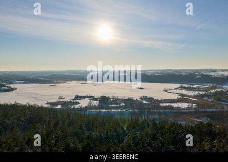 Aus der Vogelperspektive auf einen gefrorenen See umgeben von schneebedeckten Feldern und dichten immergrünen Wäldern unter der hellen Wintersonne in Litauen. Stockfoto