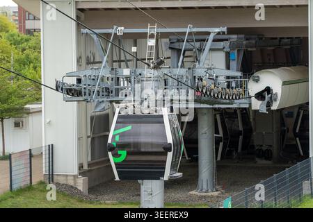 Seilbahn, die unter einer Metallkonstruktion aufgehängt ist. Dunkles Fahrerhaus mit grünen Markierungen sichtbar. Industrielle Kulisse mit Maschinen und Drähten. Keine Personen vorher Stockfoto