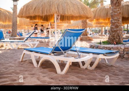 Leere blaue Sonnenliegen am Sandstrand unter Strohschirmen in einem tropischen Badeort tagsüber. Sommertourismus und Urlaubsleben. Stockfoto