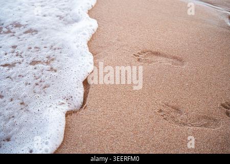 Meereswelle mit weißem Schaum, der bei Sonnenuntergang über Sandstrand mit menschlichen Spuren in der Nähe der Küste gespült wird. Sommerreisen und Natur an der Küste. Stockfoto