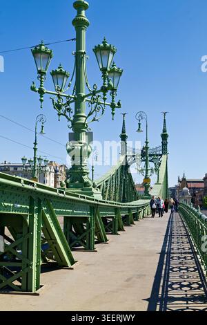 An einem sonnigen Tag spazieren die Menschen entlang der Freiheitsbrücke in Budapest Ungarn. Die Brücke verfügt über eine grüne Metallkonstruktion und Straßenlaternen. Stockfoto