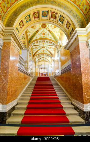 Ein roter Teppich läuft die Treppe hinauf in der Ungarischen Staatsoper. Die Wände sind mit Kunstwerken und kunstvollen Mustern versehen. Lichter erhellen den Raum. Stockfoto