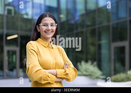 Junge professionelle Frau, die selbstbewusst lächelt, während sie mit überkreuzten Armen vor einem modernen Glasbürogebäude steht und Erfolg, Führung und Professionalität des Unternehmens symbolisiert Stockfoto