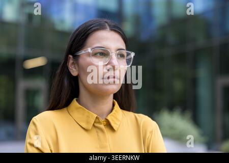 Junge Geschäftsfrau in gelbem Hemd und klarer Brille, die selbstbewusst vor einem modernen Bürogebäude steht und Ehrgeiz, Fokus und städtische berufliche Entschlossenheit projiziert Stockfoto
