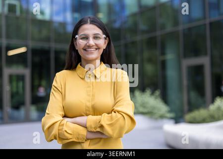Eine junge Berufsfrau mit Brille, die ein gelbes Hemd trägt, steht draußen vor einem modernen Firmengebäude und lächelt selbstbewusst mit gekreuzten Armen Stockfoto